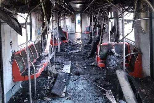 Interior de um vagão de trem completamente destruído por um incêndio. Bancos vermelhos estão queimados e deformados, o teto está parcialmente derretido com fios expostos pendendo, e o piso está coberto de fuligem e destroços. Ao fundo, uma porta aberta deixa entrar luz natural, destacando a extensão dos danos.