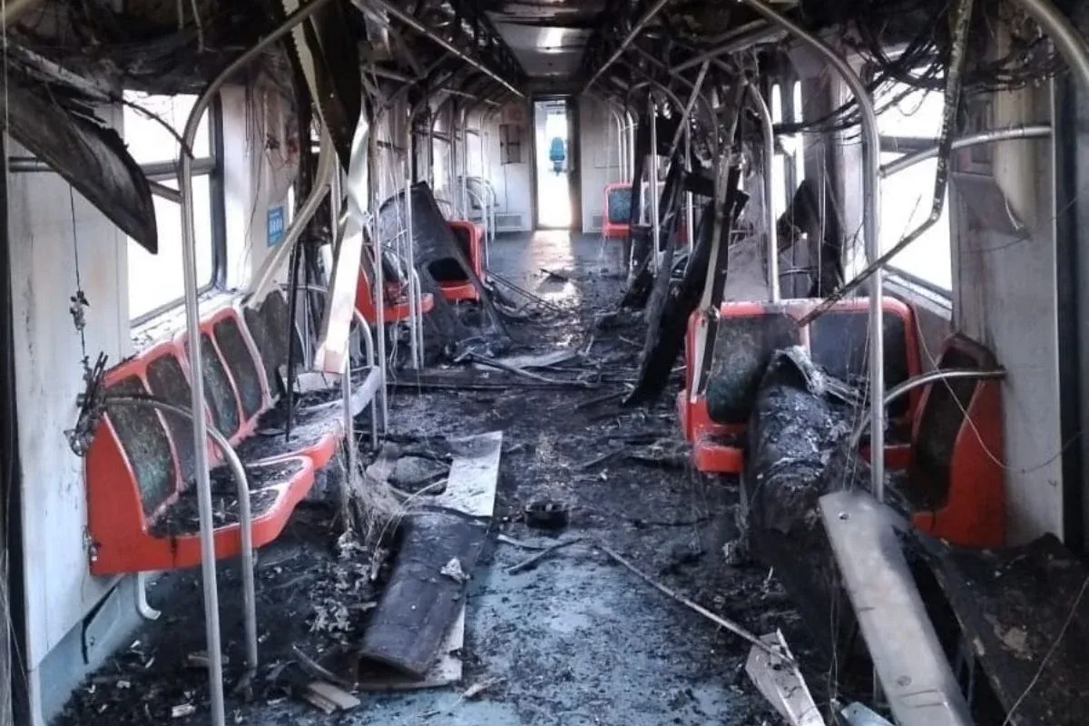 Interior de um vagão de trem completamente destruído por um incêndio. Bancos vermelhos estão queimados e deformados, o teto está parcialmente derretido com fios expostos pendendo, e o piso está coberto de fuligem e destroços. Ao fundo, uma porta aberta deixa entrar luz natural, destacando a extensão dos danos.