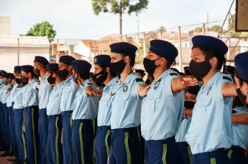 Estudantes de uma escola cívico-militar estão enfileirados, usando uniformes azul-claros, calças e boinas azuis e máscaras pretas. Alguns mantêm o braço direito estendido à frente durante uma formação.