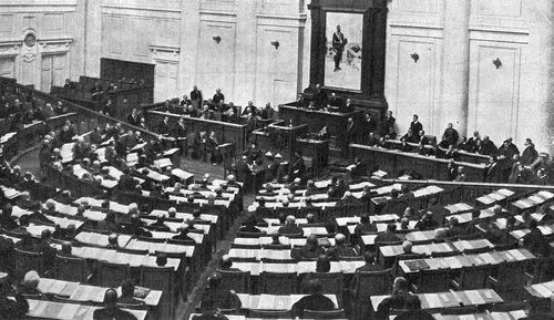 Fotografia histórica em preto e branco mostra uma sessão da Duma no Palácio Táuride, com dezenas de deputados sentados em fileiras semicirculares enquanto um parlamentar discursa diante da assembleia.