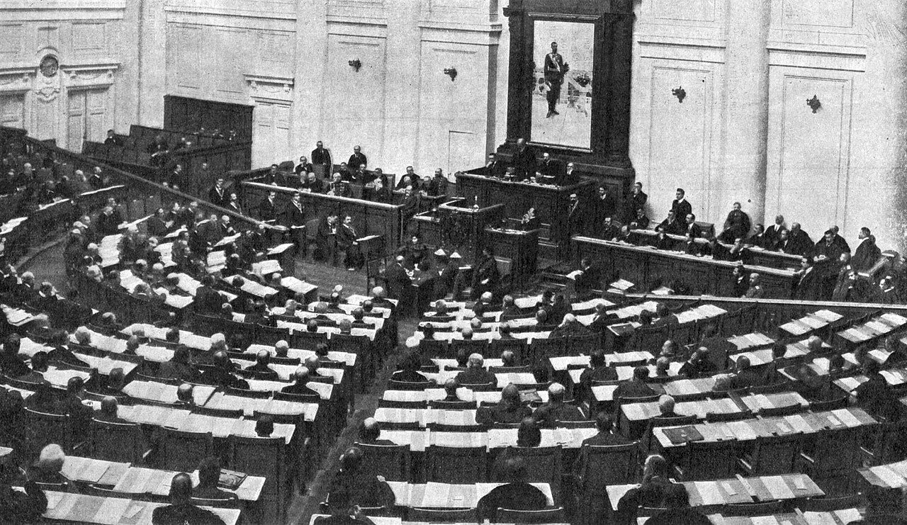 Fotografia histórica em preto e branco mostra uma sessão da Duma no Palácio Táuride, com dezenas de deputados sentados em fileiras semicirculares enquanto um parlamentar discursa diante da assembleia.