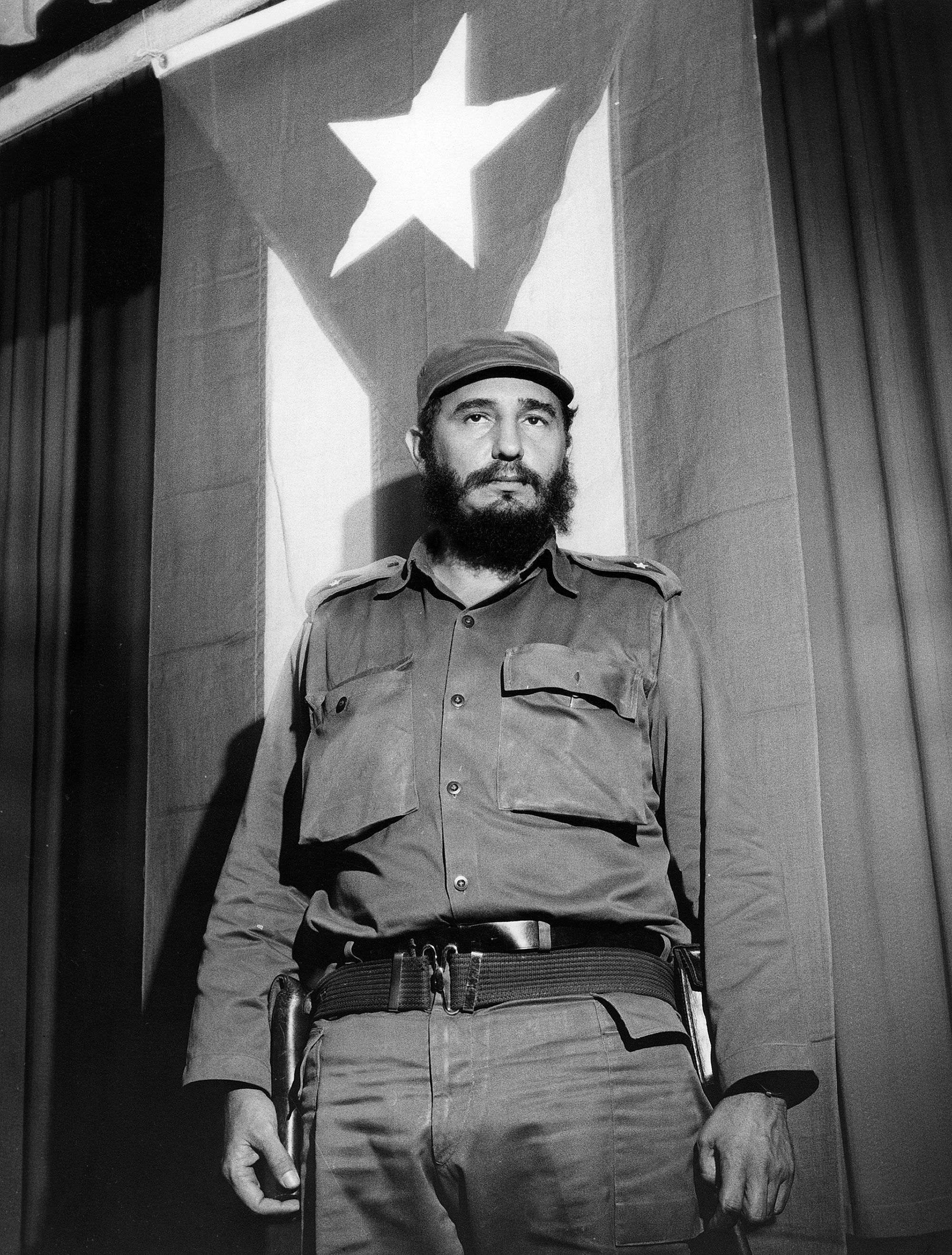 Fotografia em preto e branco de Fidel Castro, de barba e uniforme militar, em pé diante de uma bandeira cubana com uma grande estrela ao fundo.