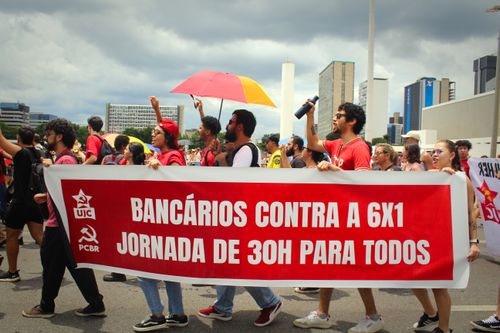 Grupo de manifestantes marcha por uma avenida segurando uma faixa vermelha com os dizeres: “Bancários contra a 6x1. Jornada de 30h para todos”. Ao fundo, prédios, céu nublado e pessoas com bandeiras e um guarda-chuva colorido.