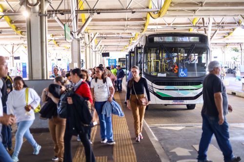 Fotografia de um terminal de ônibus urbano coberto, movimentado por dezenas de pessoas caminhando pela plataforma. Ao centro, um ônibus branco com detalhes em verde está parado para embarque e desembarque. Na parte frontal do veículo há um símbolo de acessibilidade para cadeirantes e um letreiro eletrônico pouco legível. Passageiros de diferentes idades circulam em várias direções, alguns carregando bolsas, mochilas ou casacos. O piso possui faixa tátil amarela para orientação de pessoas com deficiência visual. A estrutura metálica do terminal, com vigas e tubulações aparentes no teto, ocupa toda a parte superior da imagem, enquanto placas de identificação das plataformas aparecem ao fundo. A cena transmite a movimentação típica de um terminal de transporte coletivo em horário de grande circulação.