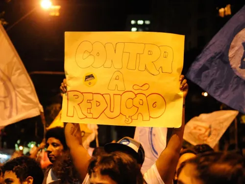 Manifestante ergue um cartaz com a frase “Contra a redução” durante ato noturno. Ao redor, outras pessoas participam da manifestação segurando bandeiras.
