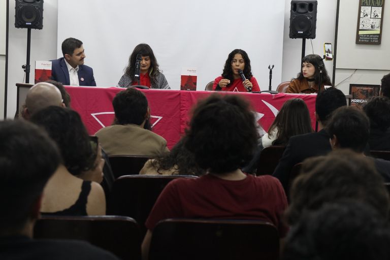 Público acompanha uma mesa de debate em um auditório. Quatro pessoas estão sentadas à frente, atrás de uma mesa coberta por uma bandeira vermelha, enquanto uma das participantes fala ao microfone.