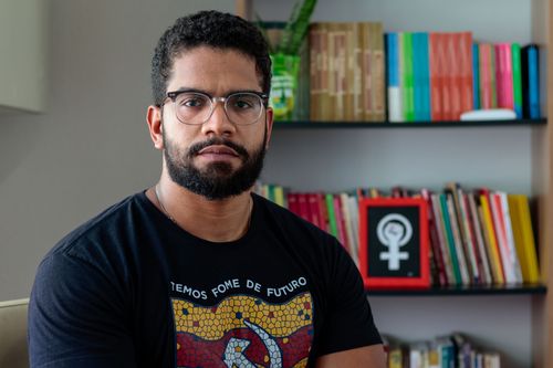 Fotografia de Jones Manoel, de óculos, barba e cabelo curto, usando camiseta preta estampada. Ele olha para a câmera com expressão neutra, tendo ao fundo uma estante com livros e uma planta.
