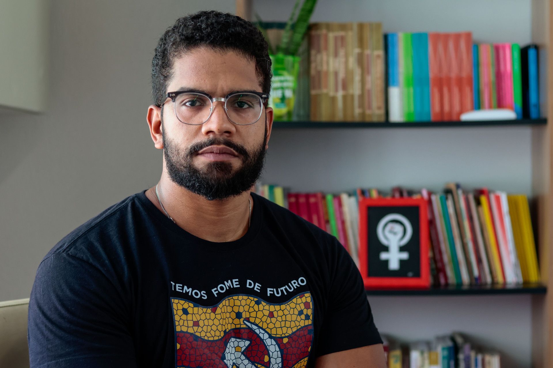 Fotografia de Jones Manoel, de óculos, barba e cabelo curto, usando camiseta preta estampada. Ele olha para a câmera com expressão neutra, tendo ao fundo uma estante com livros e uma planta.